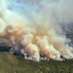 Uma silhueta de duas crianças em um balanço e duas pessoas que ficam para o lado direito, enquanto o sol à distância um vermelho brilhante brilha no meio de uma névoa de fumaça.