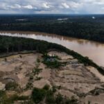 Uma área enlameada é mostrada, com três pessoas no meio dela. As árvores são visíveis além delas, com um rio atrás dele.