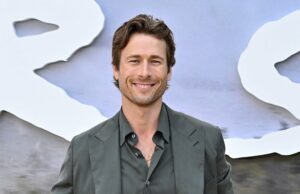 Glen Powell É uma estrela no filme de bombeiros de Ron Howard, Amazon MGM Uma mulher com cabelos ruivos usando uma camiseta branca e jeans fica perto de um homem com uma loira, flui cabelos em um moletom azul relaxado e jeans. Tem uma mão na cintura e eles ficam em frente ao fundo, sugerindo uma pequena empresa na cidade