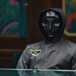 Homem sangrento e intenso em roupas formais fica abaixo da iluminação fria, seu smoking marcado com