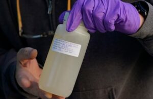 Alberta Waste Water desbloqueou informações importantes sobre o Covid-19. Também pode ajudar com o sarampo? Uma mulher com cabelos pretos e óculos se inclina para a frente e aponta para uma tela de fonte de água do banheiro. Um prato na parede atrás de leituras,