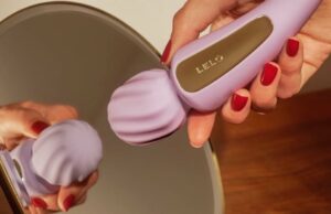 Lelo acaba de lançar um vibrador mágico de dupla ponta ultra chique Uma imagem de dois panela do vibrador de troca lilás lelo. À esquerda, o brinquedo repousa em um pano de veludo verde escuro. No lado direito, uma mão com unhas vermelhas segura o vibrador em um espelho de mesa, que mostra sua reflexão.