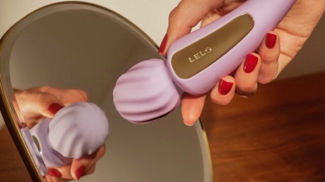 Uma imagem de dois panela do vibrador de troca lilás lelo. À esquerda, o brinquedo repousa em um pano de veludo verde escuro. No lado direito, uma mão com unhas vermelhas segura o vibrador em um espelho de mesa, que mostra sua reflexão.