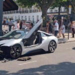 Silver BMW i8 com portas abertas após uma colisão, cercada por observadores.