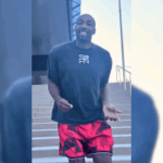 Gilbert Arenas dança da prisão e nega o envolvimento após a prisão ilegal do poker. Gilbert Arenas ri e gesticula com as mãos enquanto ele termina uma escada em roupas informais, uma camiseta preta e shorts vermelhos, do lado de fora de um prédio que parece ser um centro de detenção.