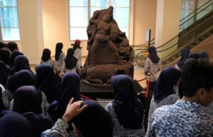 A Holanda retorna ‘Java Guy’, inúmeros fósseis da era colonial extraídos da Indonésia Três mulheres são visíveis em primeiro plano embaçado, com uma estátua de pedra ao fundo. O status está em foco e iluminado por um holofote.