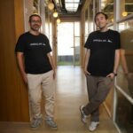 The two co-founders stand abreast in an office hallway wearing black logo t-shirts.