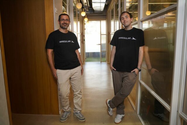 The two co-founders stand abreast in an office hallway wearing black logo t-shirts.