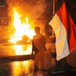 Um homem carrega a bandeira nacional indonésia quando um cargo de polícia de trânsito é queimado durante um protesto após a morte de um piloto de entrega em confrontos entre policiais e estudantes que protestam contra os legisladores, fora da sede da polícia regional em Jacarta, Indonésia, sexta -feira, 29 de agosto de 2025. (AP Photo/Tatan Syuflana)