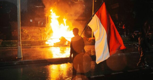 Um homem carrega a bandeira nacional indonésia quando um cargo de polícia de trânsito é queimado durante um protesto após a morte de um piloto de entrega em confrontos entre policiais e estudantes que protestam contra os legisladores, fora da sede da polícia regional em Jacarta, Indonésia, sexta -feira, 29 de agosto de 2025. (AP Photo/Tatan Syuflana)