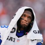 Ceedee Lamb em uma camisa branca do Dallas Cowboys com uma toalha sobre a cabeça, caminhando pela linha lateral durante um jogo.