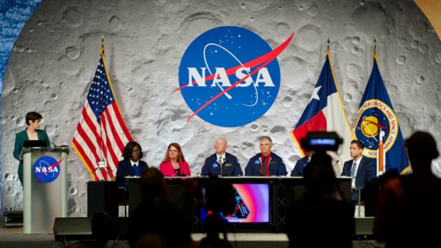 Um painel de seis pessoas se senta no palco em um evento da NASA, emoldurado pelo logotipo da NASA em um cenário da lua, com bandeiras dos EUA e do Texas ao lado deles.