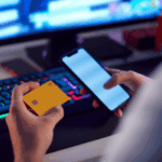 GameBleAware compartilha preocupação como os números de danos ao jogo dobram. Pessoa segurando um cartão de crédito amarelo e um smartphone na frente de uma configuração de computador com um teclado e monitor de retroilumação colorida, sugerindo pagamento on -line ou atividade de transação digital.