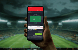 Inserir informações atributos de sportsbook novíssimos para melhorar a experiência do cliente A ENTAin apresentou uma série de novos recursos como parte de sua 'estratégia de inovação de produtos' para aprimorar suas ofertas de apostas nos Estados Unidos, no Reino Unido e no Brasil. Imagem decorativa de uma pessoa segurando um telefone na frente de uma imagem turva de um tom de estádio.