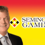 O COO da Seminole Gaming fala contra o aumento do jogo ilegal na Flórida. David Hoenemeyer em um terno preto, camisa branca e gravata cinza, sorrindo. Logotipo de jogos seminole ao lado dele em cima do fundo laranja e amarelo.