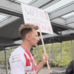 O homem estava usando um top de futebol alemão e segurando uma placa, com uma pessoa ao lado dele com o rosto embaçado.