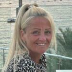 Anita Rose está vestindo um topo de estampa de animais e sentou -se em frente a uma grade à beira do mar. Seu cabelo loiro está em um rabo de cavalo alto e ela está olhando diretamente para a câmera e sorrindo. No fundo é uma palmeira e o mar.