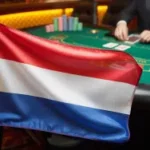 Uma fotografia da bandeira da Holanda exibida com destaque em primeiro plano, com suas distintas listras horizontais de tecido vermelho, branco e azul ondulando suavemente. A bandeira é posicionada em um suporte ou mantida em ângulo, criando profundidade e atraindo a atenção do observador para suas cores vibrantes. Atrás da bandeira, ligeiramente fora de foco, é visível uma mesa de jogo de cassino com superfície de feltro verde, com fichas de pôquer espalhadas por ela. Dutch Ksa alerta sobre crescimento do mercado ilegal de jogos de azar online