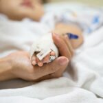 A mão de uma mãe segurando a mão de um bebê, que possui um tubo IV médico anexado.