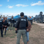 A operação do FBI em Idaho leva à acusação do grande júri de cinco pessoas em um caso de jogo. Policiais vestindo coletes de “Oficial de Liberdade Condicional do Estado de Idaho” detêm um indivíduo durante uma operação ao ar livre. Vários agentes e veículos, incluindo um camião blindado, são visíveis num campo rural sob um céu parcialmente nublado.