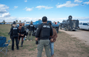 A operação do FBI em Idaho resulta em uma grande acusação judicial de 5 em situação de apostas A operação do FBI em Idaho leva à acusação do grande júri de cinco pessoas em um caso de jogo. Policiais vestindo coletes de “Oficial de Liberdade Condicional do Estado de Idaho” detêm um indivíduo durante uma operação ao ar livre. Vários agentes e veículos, incluindo um camião blindado, são visíveis num campo rural sob um céu parcialmente nublado.