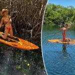 Brie Larson se ajoelhando em um paddle de laranja em um corpo de água com árvores verdes exuberantes ao fundo.