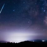 Estrelas da Via Láctea com trilhas de chuva de meteoros e silhuetas rurais.