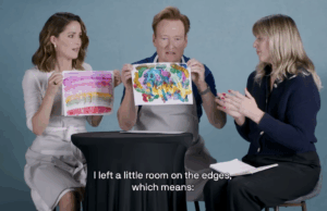 Conan OBrien e Rose Byrne fazendo tratamento artístico saem alegremente dos trilhos Três pessoas sentam-se atrás de uma mesa coberta com materiais de arte. Duas das pessoas, um homem e uma mulher, seguram pinturas.