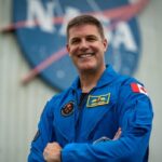 Quatro astronautas em macacões azuis estão sentados em um palco e viram para olhar para um vídeo de si mesmos em trajes espaciais de laranja.