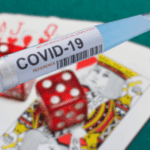 Fraudador de empréstimos COVID condenado por usar fundos em jogos de azar e criptografia. Um tubo de ensaio COVID-19 rotulado “COVID-19” colocado em cartas de baralho e dados vermelhos, simbolizando o uso indevido de fundos de ajuda à pandemia para jogos de azar.