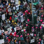 Pessoas protestam na Times Square contra as políticas de Trump, segurando vários cartazes, incluindo