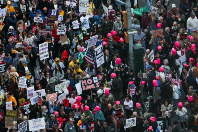 Pessoas protestam na Times Square contra as políticas de Trump, segurando vários cartazes, incluindo 
