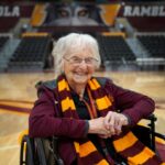 Irmã Jean Dolores Schmidt, capelã de basquete masculino da Universidade Loyola, sentada em uma cadeira de rodas em uma quadra de basquete, sorrindo para a câmera.