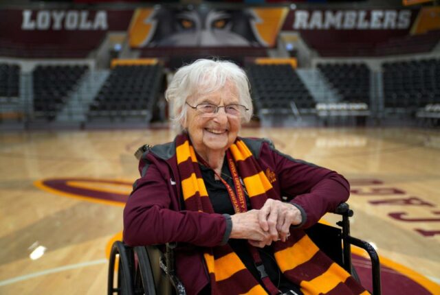 Irmã Jean Dolores Schmidt, capelã de basquete masculino da Universidade Loyola, sentada em uma cadeira de rodas em uma quadra de basquete, sorrindo para a câmera.