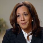 Kamala Harris olhando ligeiramente para a esquerda e sorrindo enquanto vestia um terno risca de giz e camisa de colarinho branco.