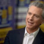 Governador da Califórnia, Gavin Newsom, vestindo terno, falando durante uma entrevista com um fundo desfocado do SEIU 2020.