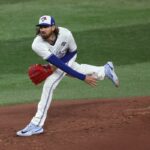 Jogador de beisebol do Blue Jays com cabelo comprido e barba, vestindo uma camisa branca, camiseta azul e um emblema da World Series na manga, segurando uma luva vermelha e olhando para frente com a boca aberta.