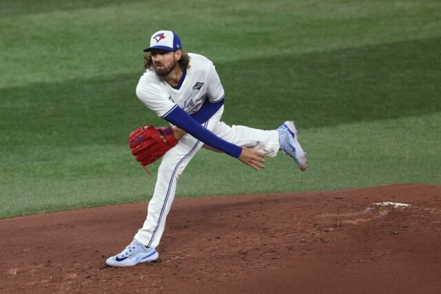 Previsão do jogo 6 de Dodgers x Blue Jays: escolhas, Jogador de beisebol do Blue Jays com cabelo comprido e barba, vestindo uma camisa branca, camiseta azul e um emblema da World Series na manga, segurando uma luva vermelha e olhando para frente com a boca aberta.