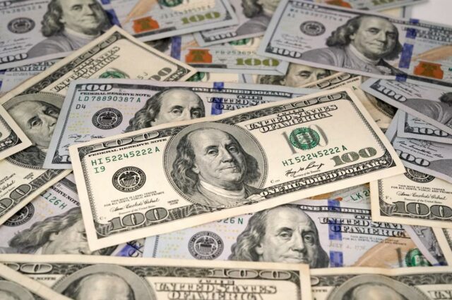 Uma jovem de cabelos castanhos cacheados, vestida com um suéter de tricô verde, tosse sentada em um sofá cinza claro.