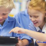Alunos usando um tablet digital em sala de aula. Há um menino e uma menina, ambos vestindo uniformes escolares e felizes e sorridentes.