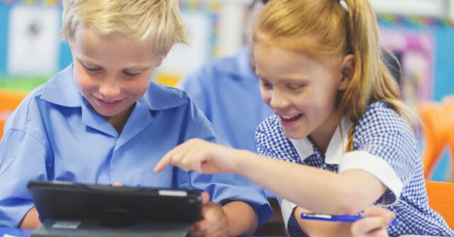 Alunos usando um tablet digital em sala de aula. Há um menino e uma menina, ambos vestindo uniformes escolares e felizes e sorridentes.