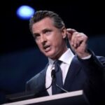 Califórnia Gavin Newsom em frente ao pódio vestindo terno. Governador da Califórnia, Newsom, criticado pelo SGLA por assinar a proibição de sorteios