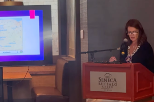 Seneca Country gera influência de praticamente US$ 2 bilhões na Mulher de óculos e blusa preta fica atrás de um pódio enquanto faz um discurso, com uma TV à sua esquerda. Nação Seneca gera impacto de quase US$ 2 bilhões na economia do oeste de Nova York