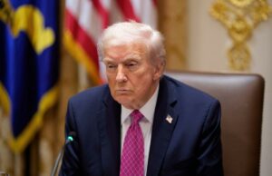 Trump anuncia aumento de tarifas de 10% sobre o Canadá enquanto a fúria sobre o ‘ato hostil’ do anúncio de Reagan atinge o ponto de ebulição Donald Trump vestindo terno escuro e gravata rosa, com distintivo de lapela com a bandeira dos EUA.