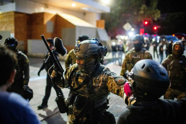 Um manifestante está envolto em uma bandeira americana observa como policiais tentam dispersar os manifestantes perto de uma instalação de imigração e fiscalização aduaneira dos EUA em Portland, Oregon