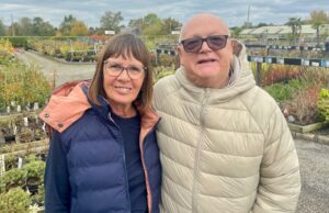 Yard centers aos 70: Por que gostamos deles? Um homem está parado em uma seção externa protegida de um centro de jardinagem. Há arbustos, estátuas e grandes plantas dispostas atrás dele sob o teto transparente sustentado por uma estrutura de metal. Ele usa óculos, camisa azul, lã cinza, casaco preto e tem cabelo grisalho curto e barba grisalha curta.