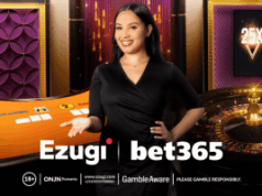 A bet365 faz parceria com a Ezugi para melhorar as ofertas de cassino ao vivo no Reino Unido, Ontário e Nova Jersey Uma sorridente dealer de cassino ao vivo está em frente a uma mesa de jogo laranja e uma roleta. Ela está usando um vestido preto e gesticula com uma das mãos em direção à mesa. O fundo apresenta painéis decorativos roxos e dourados e um gráfico multiplicador “25X” em uma tela. Os logotipos da Ezugi e da bet365 aparecem na parte inferior, junto com avisos de jogo responsável. A bet365 faz parceria com a Ezugi para melhorar as ofertas de cassino ao vivo no Reino Unido, Ontário e Nova Jersey