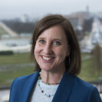 Membros do Congresso e funcionários estão na Câmara da Câmara dos EUA perto de um grande retrato de George Washington. Nancy Pelosi está perto do centro do grupo, vestindo um terno branco, cercada por assessores e legisladores. As pessoas estão sentadas em primeiro plano enquanto a câmara se enche de membros.