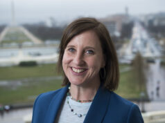 A mulher que fez história com Nancy Pelosi Membros do Congresso e funcionários estão na Câmara da Câmara dos EUA perto de um grande retrato de George Washington. Nancy Pelosi está perto do centro do grupo, vestindo um terno branco, cercada por assessores e legisladores. As pessoas estão sentadas em primeiro plano enquanto a câmara se enche de membros.