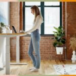 Cadeira de escritório cinza claro com braços e base cromada com rodas ao lado de uma mesa branca.